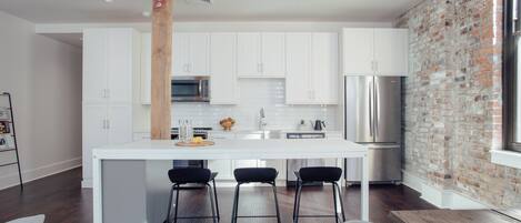 A modern kitchen with white cabinets, a central island with stools, a brick accent wall, and a stainless steel refrigerator.