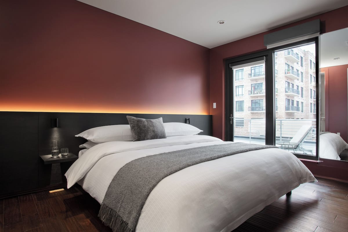 A modern hotel room with a large bed, a nightstand, a window with a view of buildings, and a dark-colored wall.