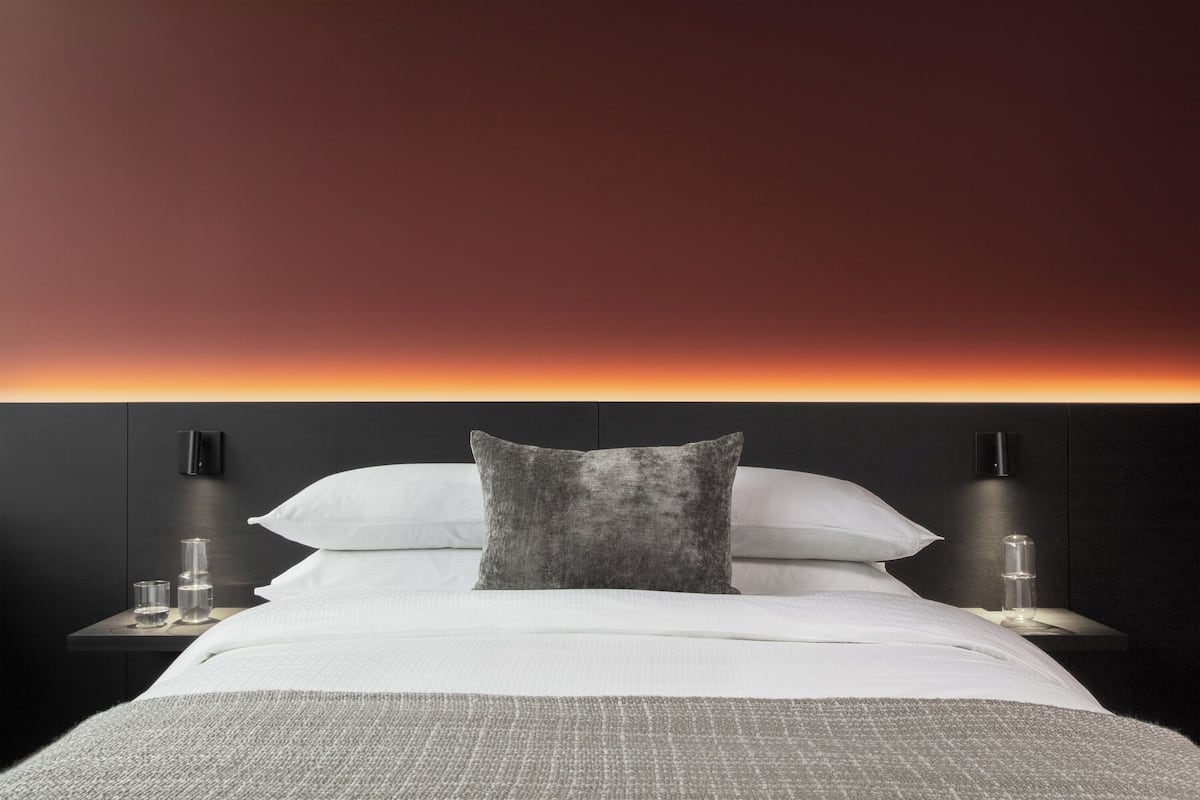 A neatly made bed with white linens and a grey pillow, flanked by two bedside tables with lamps, against a dark wall with a subtle gradient.