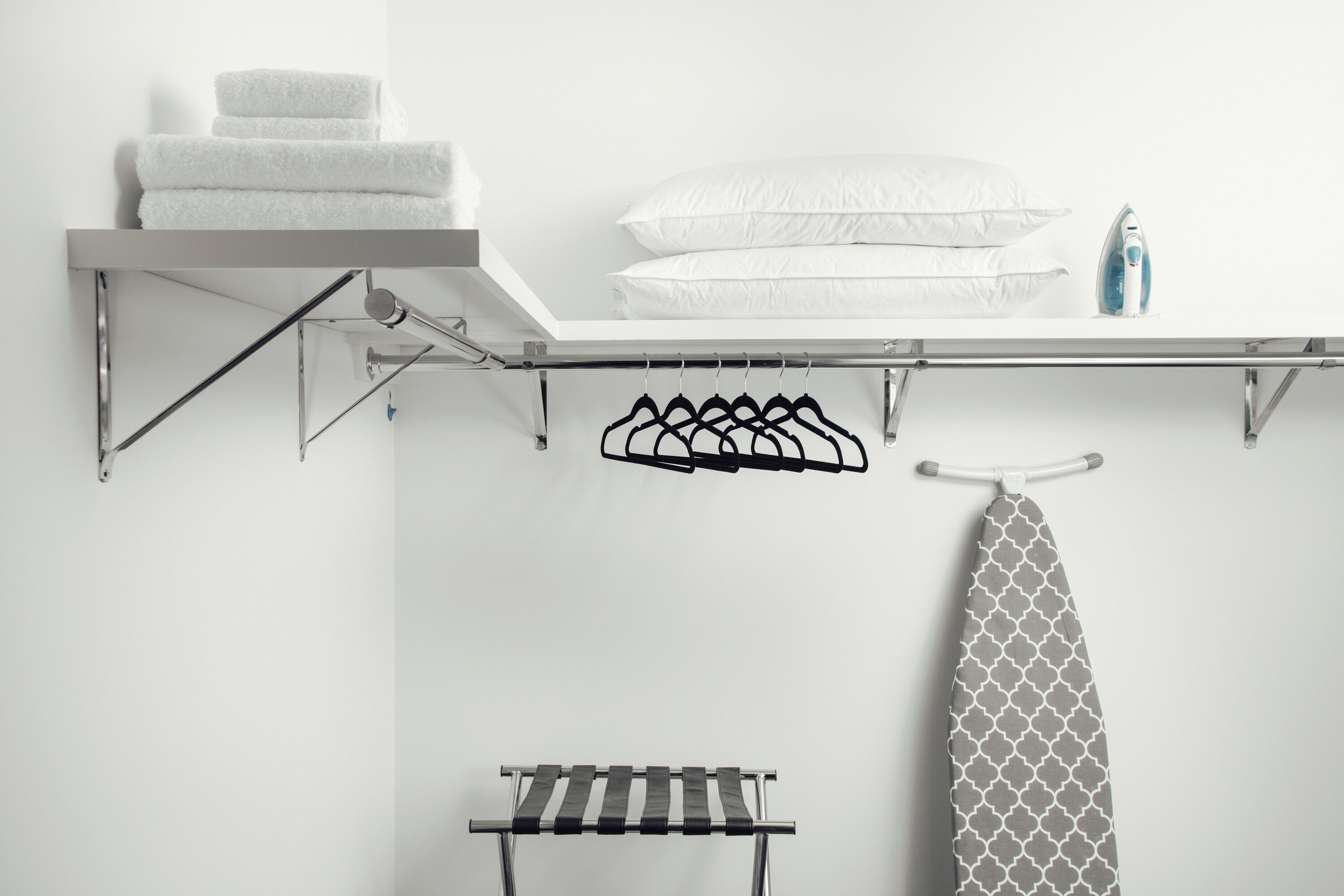 Een netjes opgeruimde badkamer met een handdoekrek, kleding die hangt, een plank met handdoeken, een hangend strijkijzer, en een handdoekhouder.