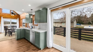 A modern kitchen with green cabinets, a white countertop, and a dining area with a table and chairs. - Conrad Vista | Fully Remodeled Waterfront Retreat (Priest River)