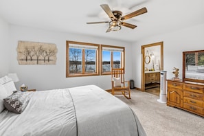 A bedroom with a bed, a wooden dresser, a chair, a ceiling fan, and a large window with a view of trees and a lake.