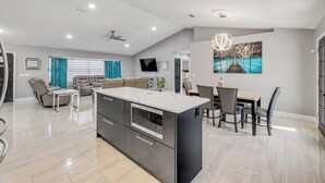 Eine moderne Küche mit Kochinsel, ein Essbereich mit Stühlen und Tisch, ein Wohnbereich mit Sofa und Fernseher sowie ein Deckenventilator.