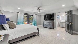 Ein modernes Schlafzimmer mit einem großen Bett, einem Deckenventilator, einem Fernseher und einem durchgehenden Türrahmen einsehbaren Badezimmer.