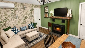 Ein Wohnzimmer mit Sofa, Hocker, Kamin, Fernseher und einem Esstisch mit Schüssel.