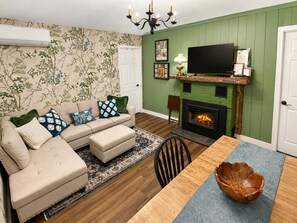 Ein Wohnzimmer mit Sofa, Hocker, Kamin, Fernseher und einem Esstisch mit Schüssel.