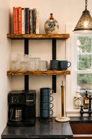Una cuina amb una cafetera, un búl, un conjunt de tasses blaves i un ram de flors decoratiu a una estant.