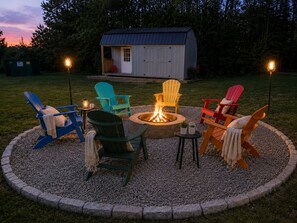 Ein runder Feuerkorb, umgeben von bunten Adirondack-Stühlen, einem kleinen Schuppen und einer Laterne.