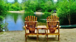 Zwei Holz-Adirondack-Stühle, die auf einen stillen See blicken, mit einem Haus im Hintergrund.