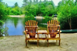 Two wooden Adirondack chairs facing a calm lake with a house in the background. - Riverside Cottage • Fireplace • Mountain Views (Bartlett)