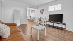 Un salon moderne avec un canapé en cuir marron, une table basse blanche, un téléviseur posé sur un meuble noir, et un coin repas à l’arrière-plan.