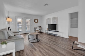 Ein modernes Wohnzimmer mit einem grauen Sofa, einem runden Couchtisch, einem Flachbildfernseher und einem großen Fenster mit Blick auf einen Balkon.