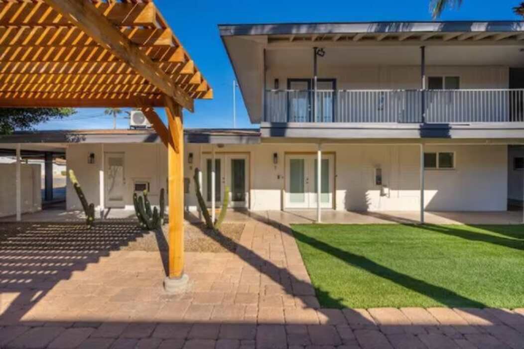 A two-story building with a flat roof, a balcony on the upper floor, and a paved courtyard with a lawn.