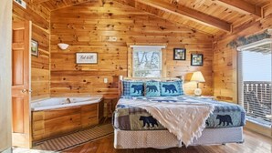 Ein gemütliches Cabin-Zimmer mit Holzvertäfelung, einem großen Bett mit Bärenmotiven, einer Badewanne und einem Fenster mit Jalousien.