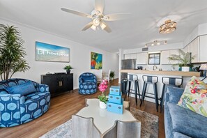 Ein modernes Wohnzimmer mit Sofa, Couchtisch und Sesseln. Es gibt eine Küchenzeile mit einer Bar und Hockern.