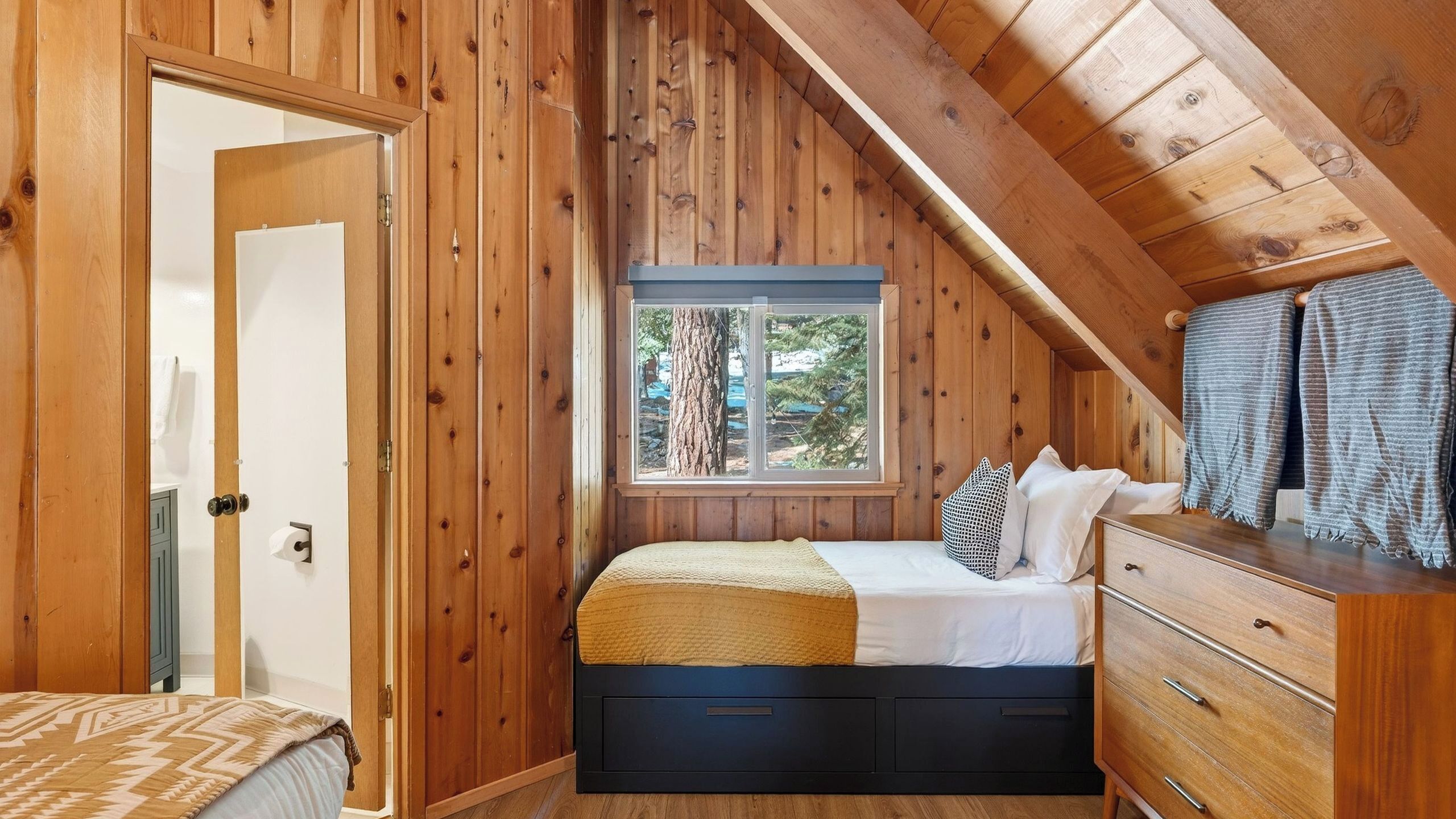 A bedroom with a wooden sloped ceiling, a window with a view of trees, a bed with a blanket, and a dresser with a mirror.