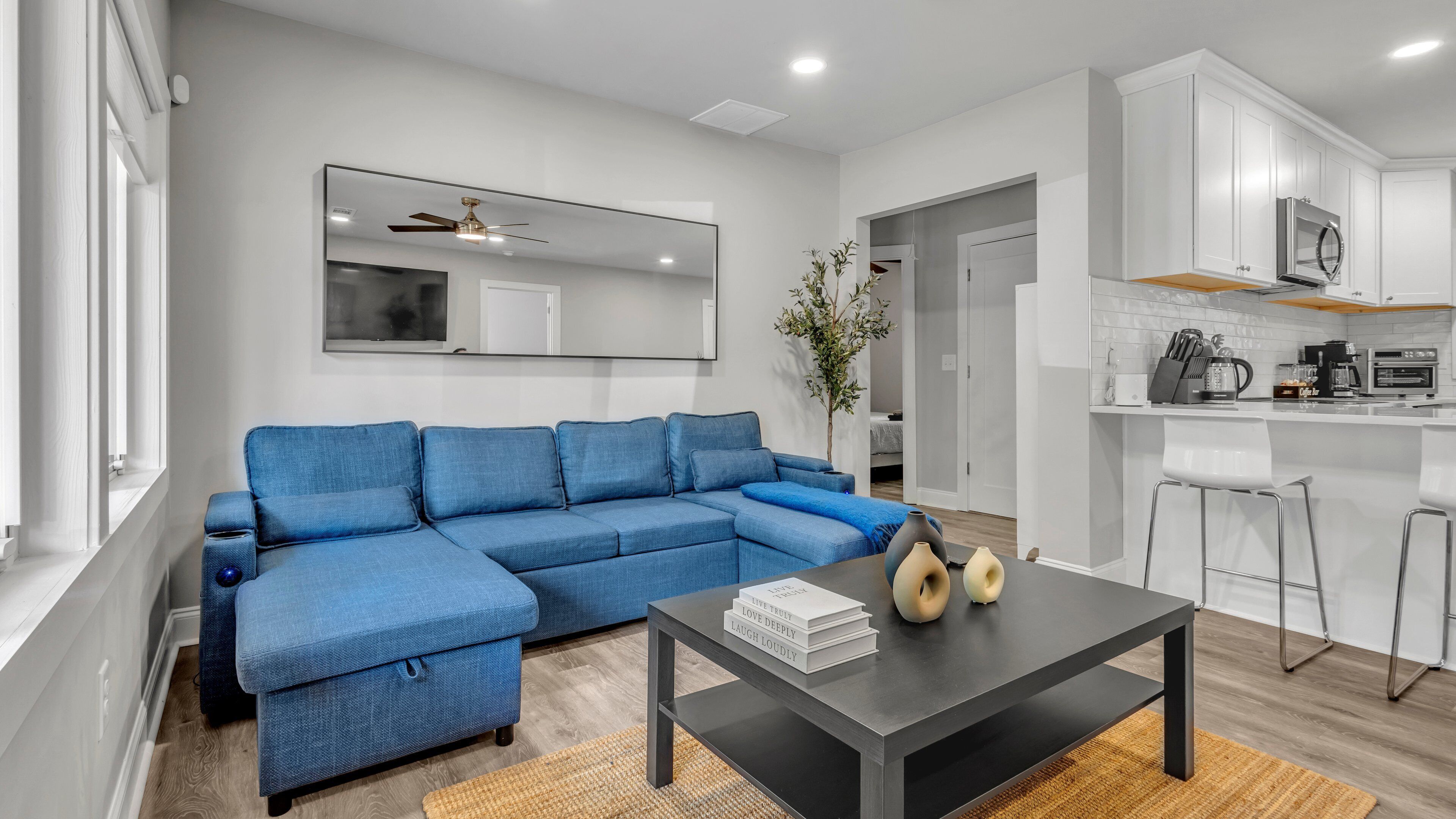 A modern living room with a sectional sofa, a coffee table, and a kitchen area with white cabinets and appliances.