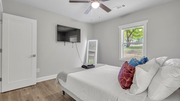 A bedroom with a bed, a TV, a ceiling fan, a window with a view, and a door.
