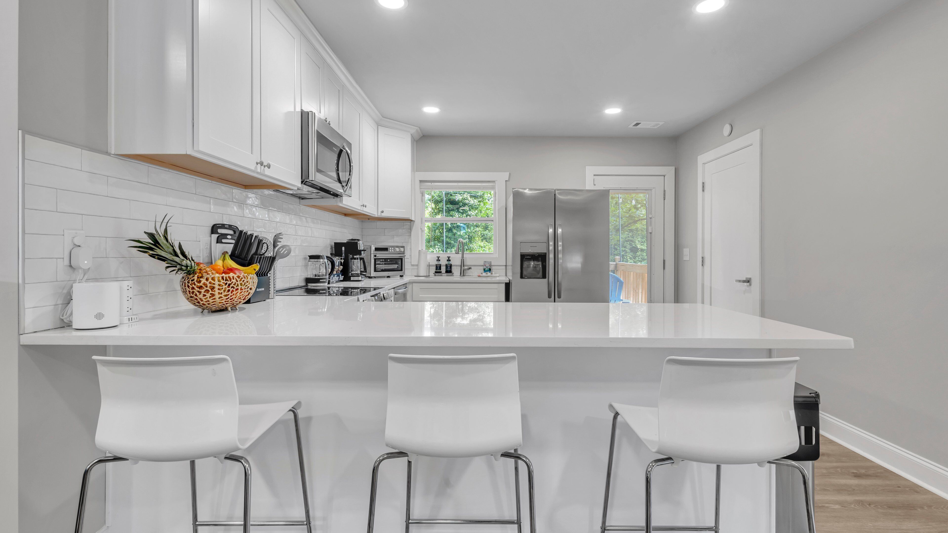 A modern kitchen with white cabinetry, a large island with bar stools, stainless steel appliances, and a view of the outdoors.
