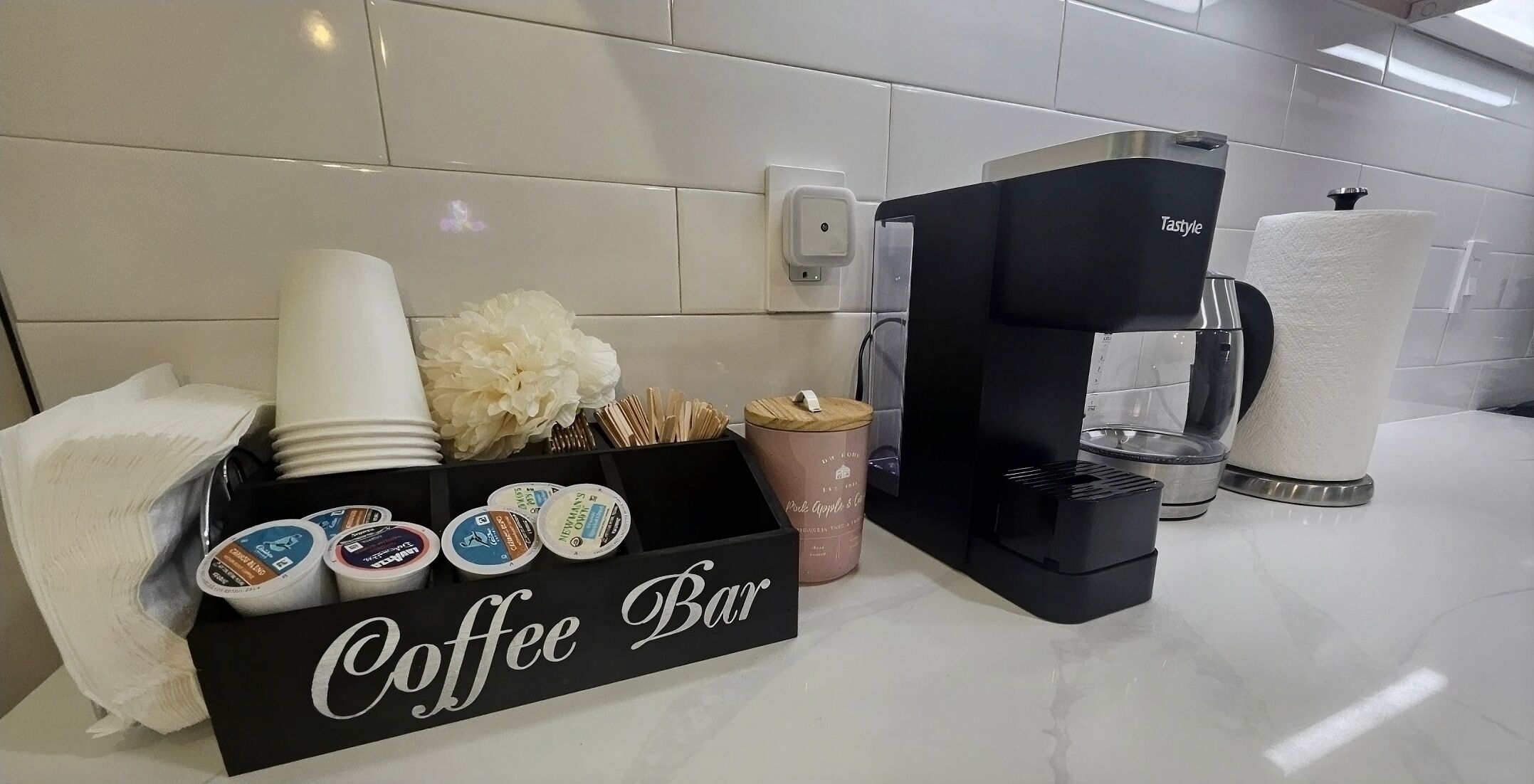 A coffee bar setup with a Tastyle coffee machine, disposable cups, a coffee mug, a box of coffee pods, and a decorative flower arrangement.