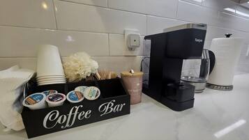 A coffee bar setup with a Tastyle coffee machine, disposable cups, a coffee mug, a box of coffee pods, and a decorative flower arrangement.