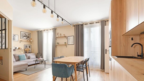 A modern living room with a sofa, coffee table, and dining table. There are curtains, shelves with decorative items, and a kitchen area with wooden cabinets and a sink. - Luminous apartement 1BDR/4P - Centre de Paris (Paris)