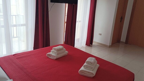 Habitación de hotel con una colcha roja, dos toallas cuidadosamente dobladas, un televisor de pantalla plana montado en la pared y vistas al exterior a través de ventanas amplias con cortinas rojas.