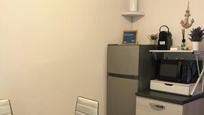 Cocina compacta con refrigerador, microondas y una pequeña encimera. Hay una mesa de comedor con sillas y un armario con cajones.