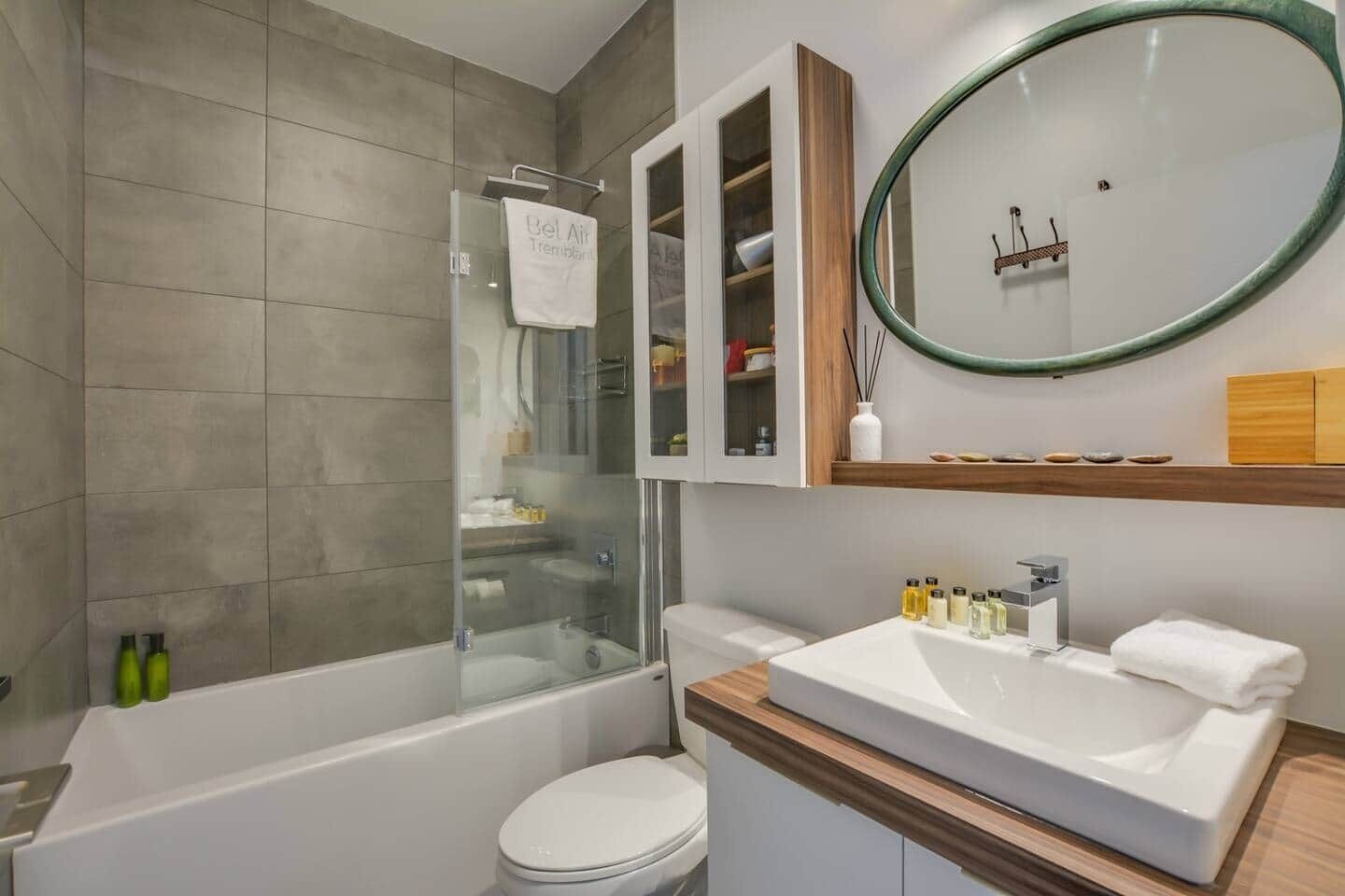 Ett modernt badrum med en duschkabin i glas, en vit toalett, ett handfat med hygienartiklar och en spegel med en träram.