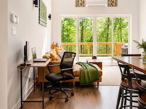 Un salon moderne comprenant un bureau en bois, un canapé confortable, une table à manger et une grande porte-fenêtre coulissante donnant sur un balcon doté d’une balustrade en bois.