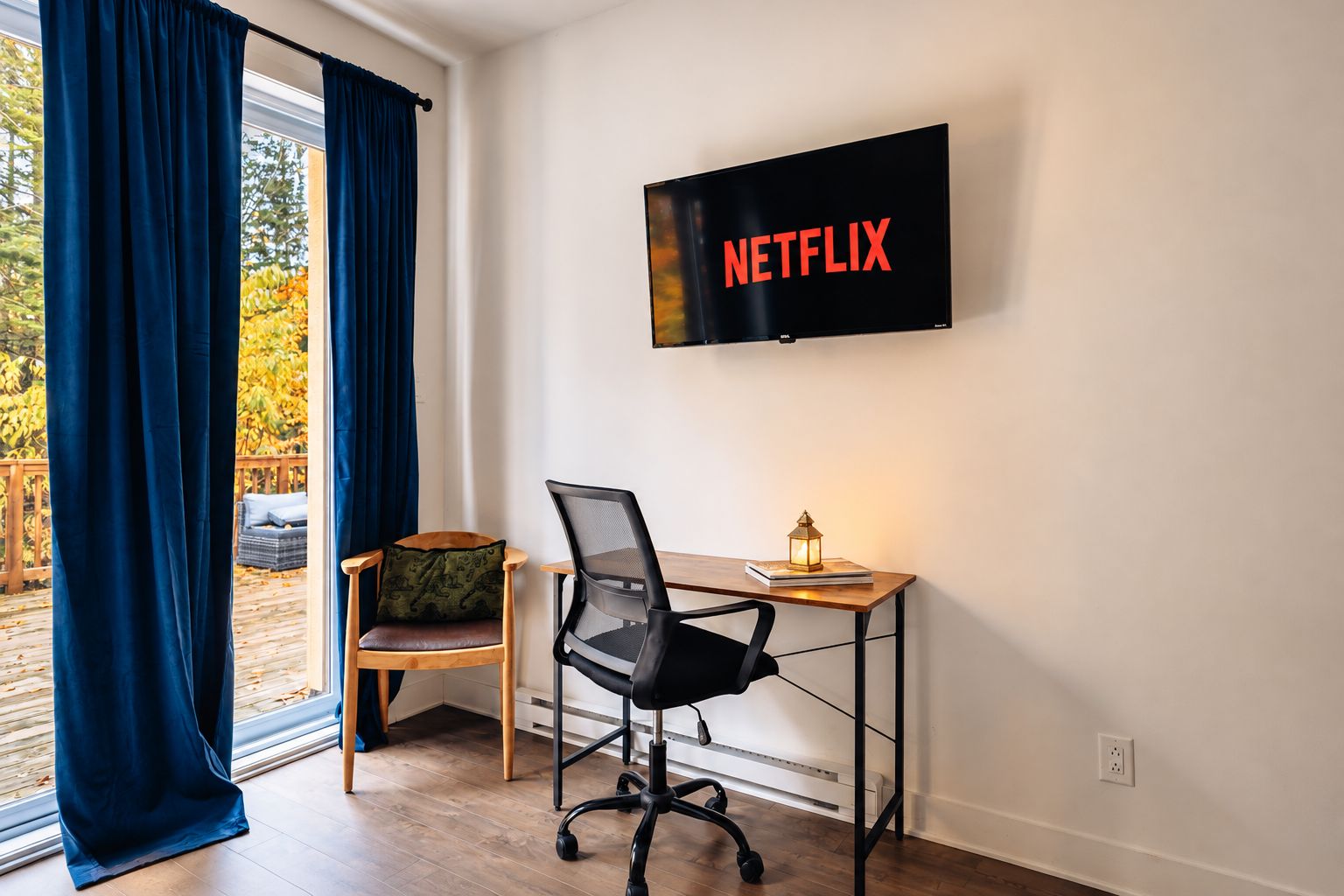 Una habitación con escritorio, silla, televisor montado en la pared mostrando 'NETFLIX' y una ventana con cortinas.