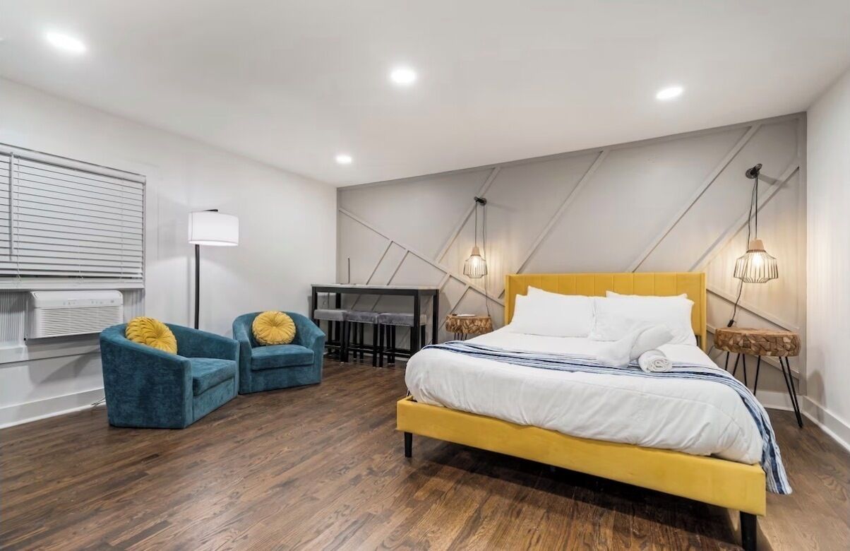 A modern bedroom with a yellow bed, two blue armchairs, a desk, and a lamp.