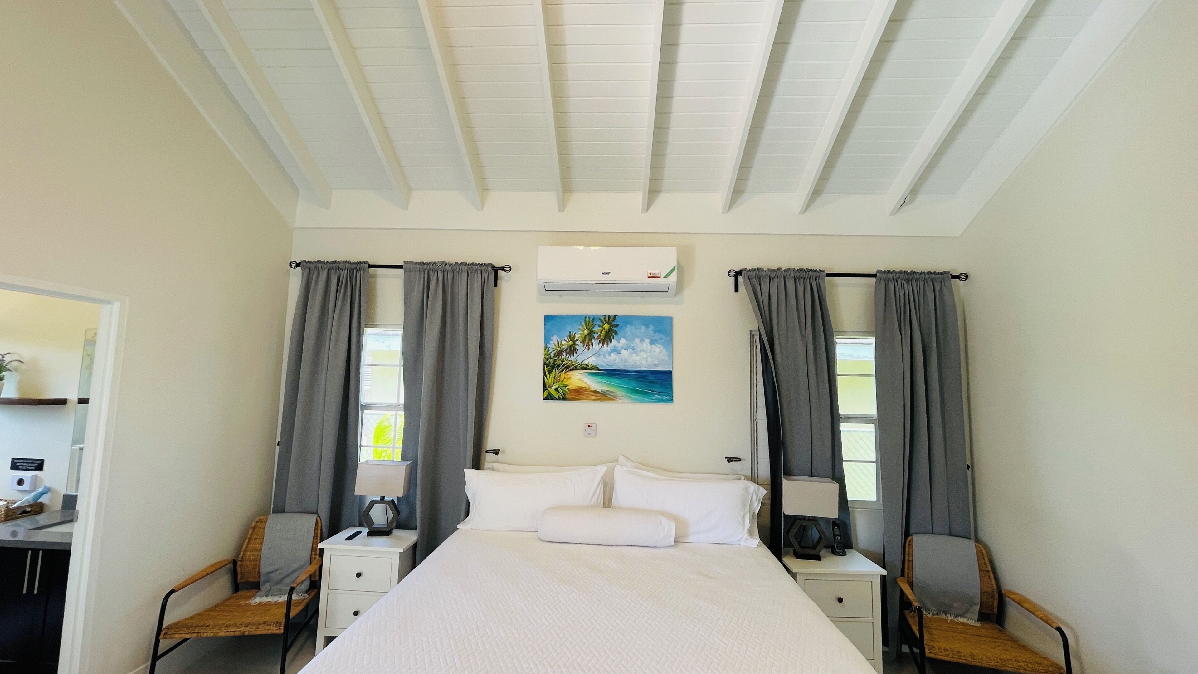 A hotel room with a bed, two bedside tables, a nightstand, a chair, a painting of a beach scene, and an air conditioning unit.