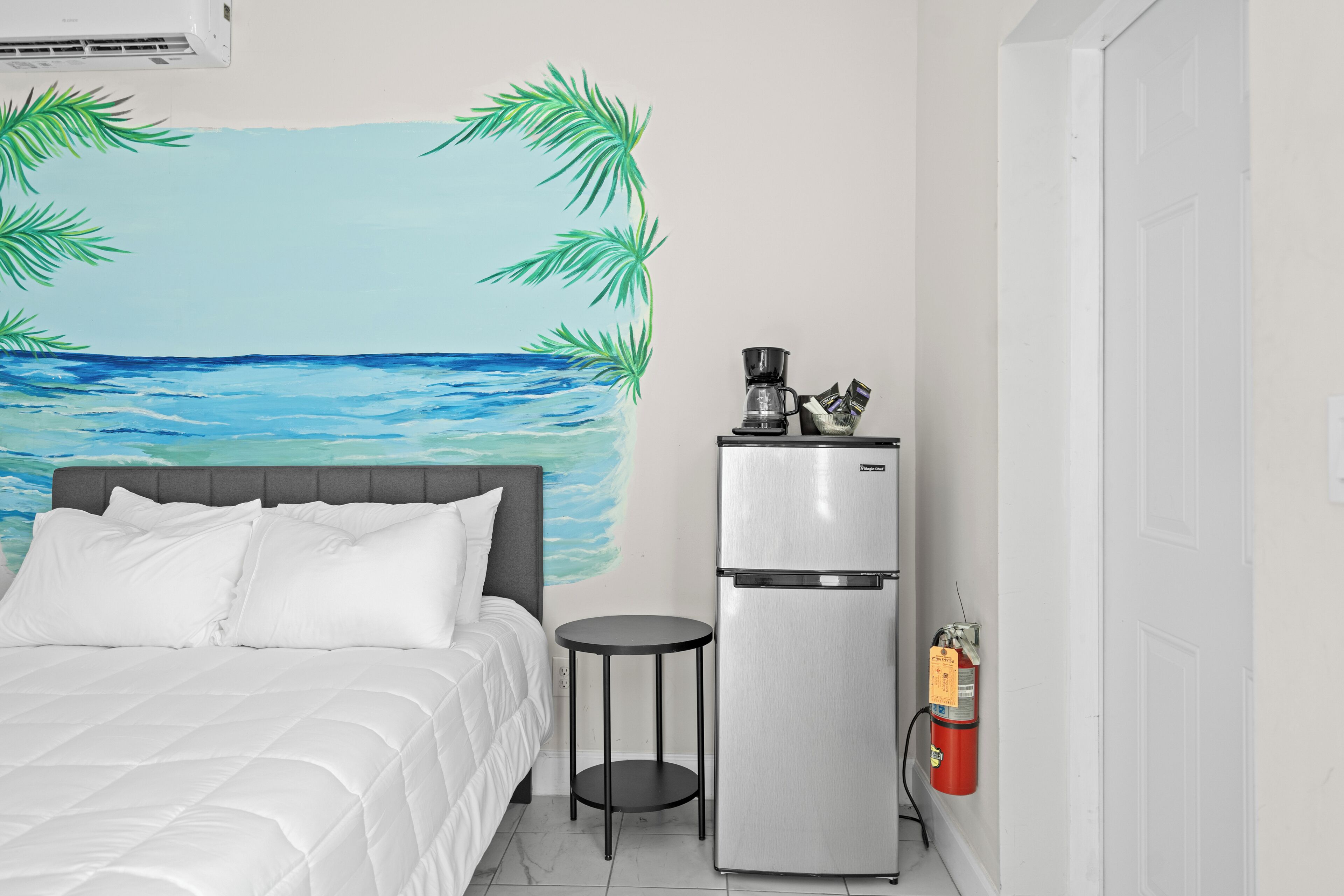 A bedroom with a bed, a small table, a refrigerator, and a wall mural of a beach scene.