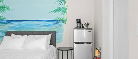 A bedroom with a bed, a small table, a refrigerator, and a wall mural of a beach scene.