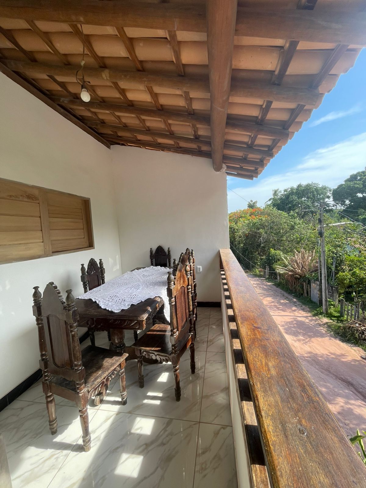 A covered patio with a wooden table and chairs, a white tablecloth, and a view of greenery.