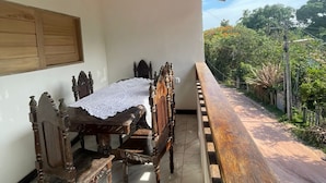 Une terrasse couverte avec une table et des chaises en bois, une nappe blanche et une vue sur la verdure.