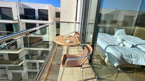 Un balcón con barandilla de vidrio, una mesa de madera con sillas y una cama con edredón blanco.