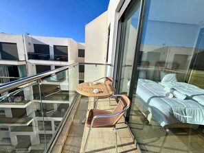 Een balkon met glazen hekwerk, een houten tafel met stoelen en een bed met een witte dekbedovertrek.