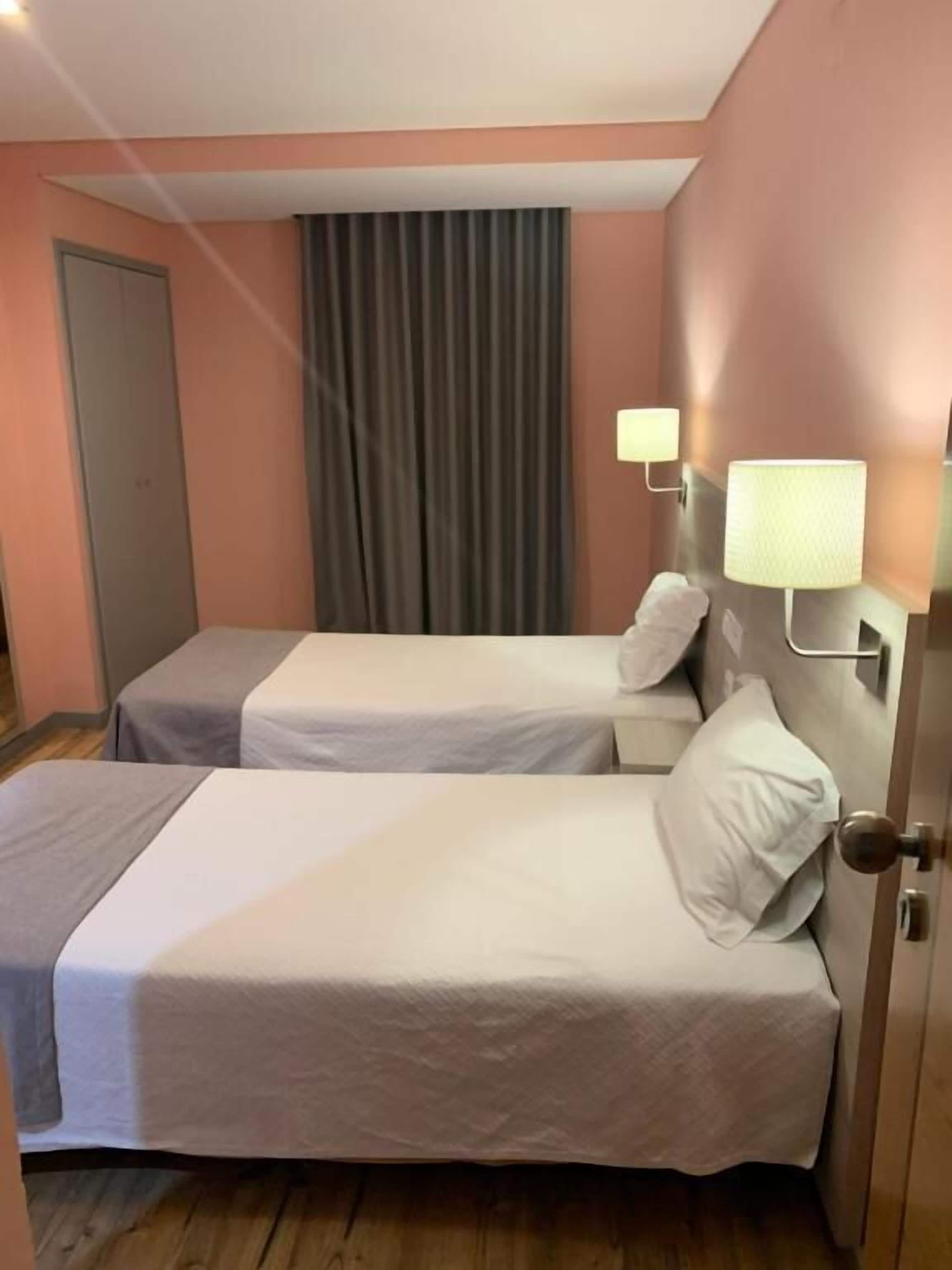 Habitación de hotel con dos camas individuales, una puerta, una cortina y dos lámparas fijadas a la pared.