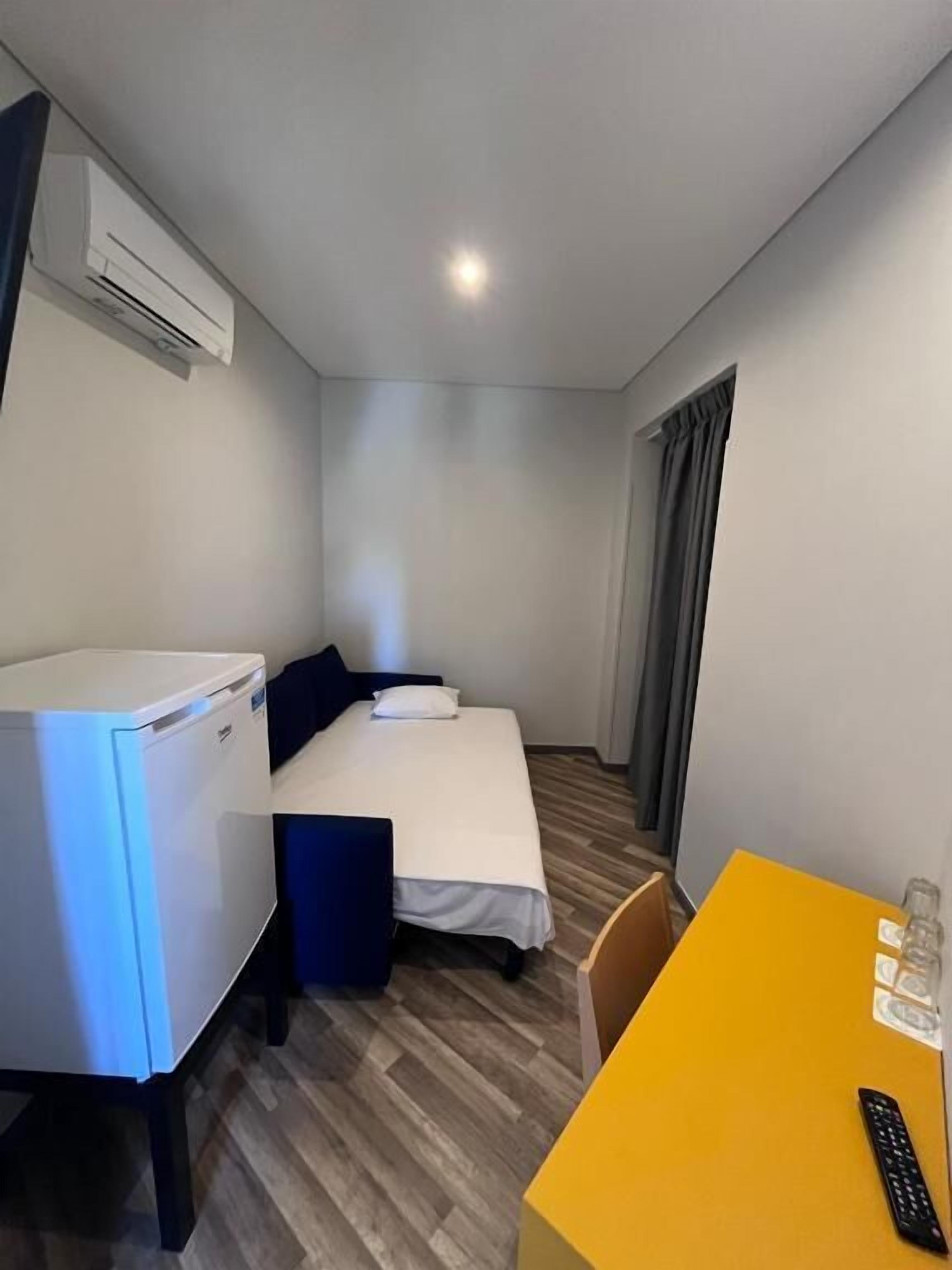 A compact hotel room with a bed, a small desk, a chair, a table, and a refrigerator.
