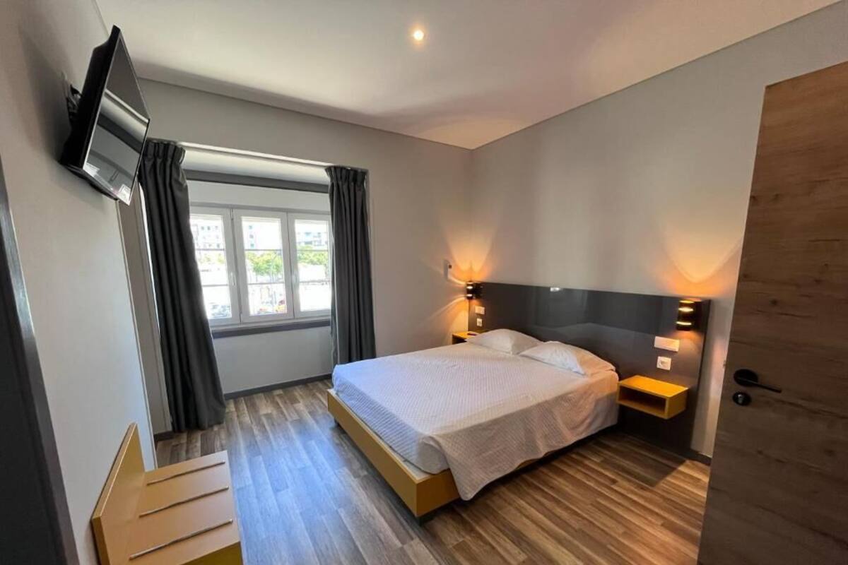 A modern hotel room with a large bed, wooden flooring, a TV mounted on the wall, and a window with curtains.