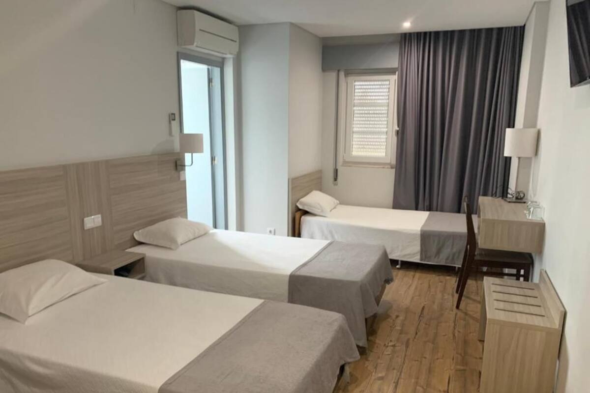 A hotel room with two single beds, a wooden floor, a window with curtains, a bedside table with a lamp, and an air conditioning unit.