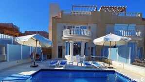 Ein Poolbereich mit Liegestühlen und Sonnenschirmen, ein Gebäude mit Balkonen und ein klarer, blauer Himmel.