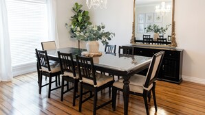 Ein Esszimmer mit einem Tisch mit Glasplatte, sechs Stühlen, einer Vase mit Grünpflanzen, einer Kandelaber und einem großen Spiegel mit goldener Rahmung.