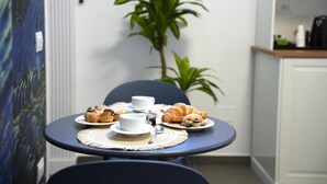 Een tafeltje met diverse gebakjes en bekers, een plantje op de achtergrond, en een keukenhoek met kasten en apparatuur.