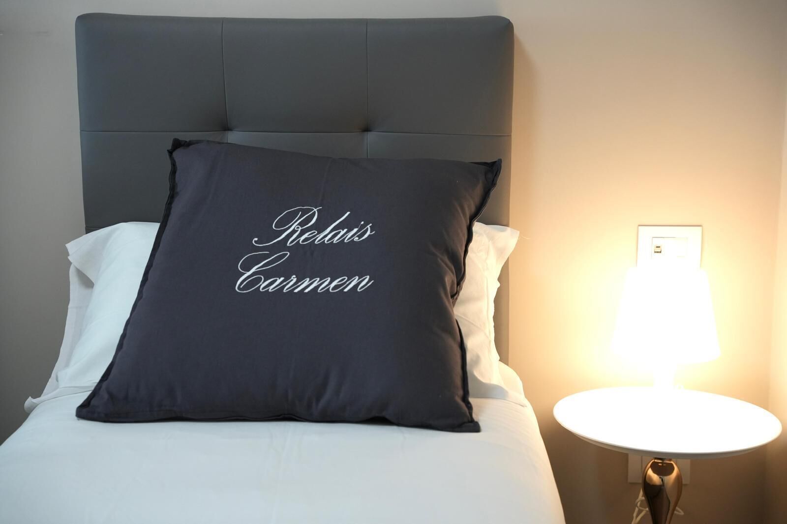 A bed with a dark gray pillow featuring the name "Redais Carmen" and a bedside lamp.