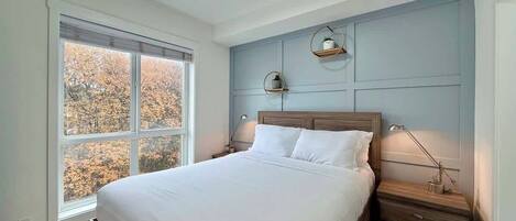 Un dormitorio con una cama grande, mesitas de noche, una ventana con vista a árboles y una lámpara en el techo.