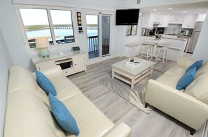 Una sala moderna con un conjunto de sofá y una mesa de centro, una cocina con alacenas blancas y un televisor de pantalla plana, y una puerta corrediza de vidrio que da a un balcón con vista a un cuerpo de agua.