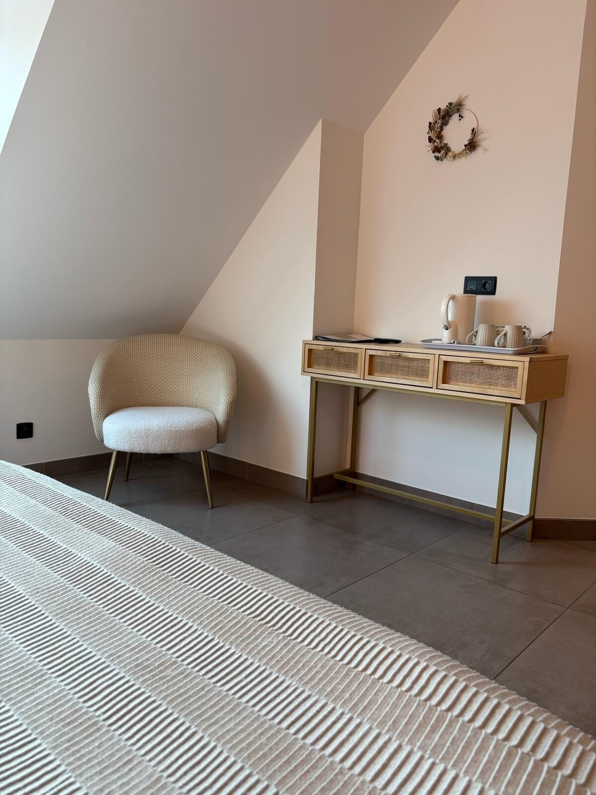 A modern bedroom with a sloped ceiling, a woven chair, a bedside table with a lamp, and a decorative wreath on the wall.